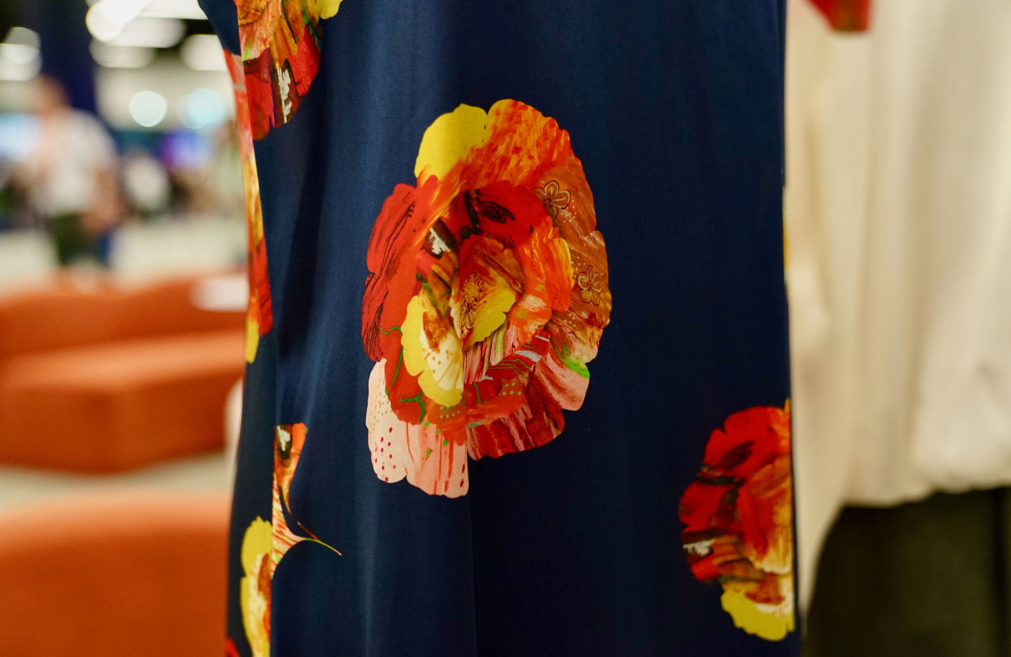 A close-up of navy blue fabric featuring large, vibrant floral patterns in red, orange, yellow, and pink tones from social enterprise, The Social Outfit. The flowers have painterly, layered petals that contrast brightly against the dark background.